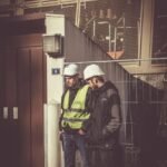 Two construction workers in safety gear standing outdoors at a building site, focused on work.